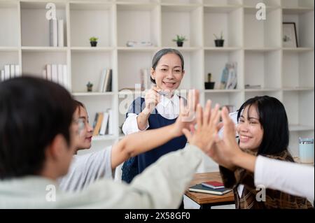 A group of cheerful Asian students putting their hands together in the classroom, brainstorming and cheering up. friendship, teamwork, education Stock Photo