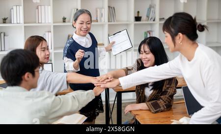 A group of cheerful Asian students putting their hands together in the classroom, brainstorming and cheering up. friendship, teamwork, education Stock Photo