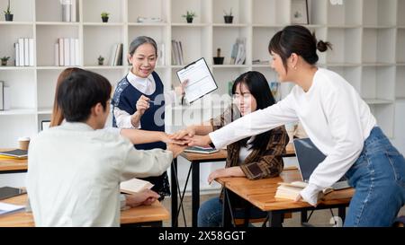 A group of cheerful Asian students putting their hands together in the classroom, brainstorming and cheering up. friendship, teamwork, education Stock Photo