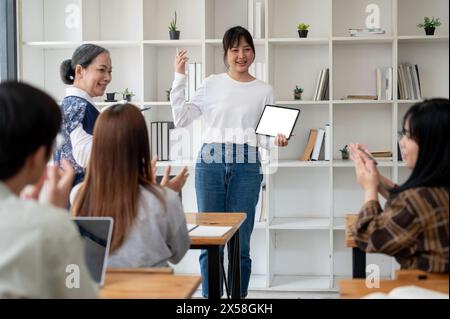 A young Asian female college student is showing her project on her digital tablet, presenting her project in the classroom to her teacher and friends. Stock Photo