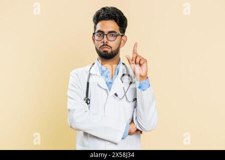 Displeased upset Indian young doctor cardiologist man reacting to unpleasant awful idea, dissatisfied with bad work, wave hand, shake head No dismiss bad idea proposal. Arabian guy on beige background Stock Photo