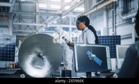 Female Engineer Uses Tablet Computer while Working on Satellite Construction. Aerospace Agency Manufacturing Facility: Scientists Build, Assemble Spacecraft for Space Exploration, Observation Mission Stock Photo