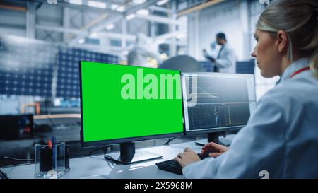 Female Engineer uses Green Screen Computer to Analyse Satellite. Aerospace Agency Manufacturing Facility: Scientists Develop, Assemble Spacecraft for Space Exploration Mission. Over Shoulder Stock Photo