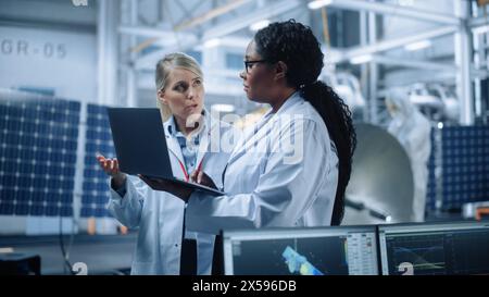 Two Female Engineers Talk, Use Computer working at Aerospace Satellite Manufacturing Facility. Diverse Team of Multi-Ethnic Scientists Doing Science Technology Research for Space Exploration Program Stock Photo