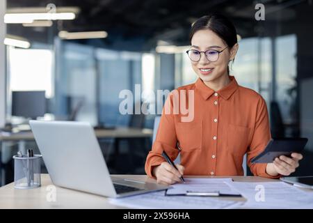 A professional businesswoman with glasses smiles while working at her office desk, using a laptop and calculator. Focus on paperwork and productivity in a corporate setting. Stock Photo