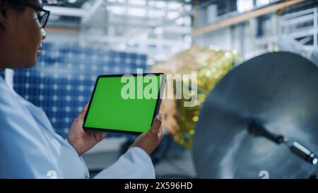 Female Engineer Uses Green Screen Digital Tablet Computer while Working on Satellite Construction. Aerospace Agency: Scientists Assemble Spacecraft for Space Exploration Mission. Over the Shoulder Stock Photo