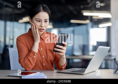 Focused Asian businesswoman in orange blouse looking puzzled while holding a smartphone at her modern office workspace. She appears to be reacting to unexpected news. Stock Photo