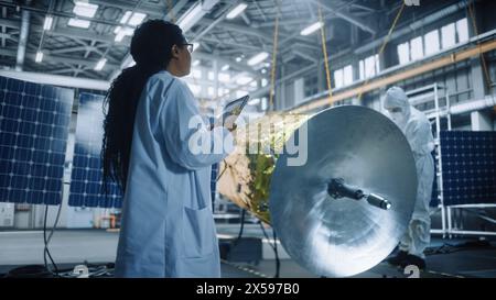 Female Engineer Uses Digital Tablet Computer while Working on Satellite Construction. Aerospace Agency: Scientist Overseeing Building, Assembly and Manufacturing of Spacecraft for Space Mission Stock Photo
