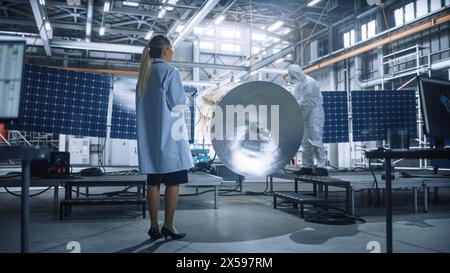 Female Engineer Uses Tablet Computer while Working on Satellite Construction. Aerospace Agency Manufacturing Facility: Scientists Build, Assemble Spacecraft for Space Exploration, Observation Mission Stock Photo