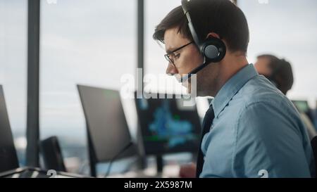 Male Air Traffic Controller with Headset Using Touchscreen Table Panel in Airport Tower. Office Room is Full of Computers with Navigation Screens, Airplane Flight Radar Data for the Team. Stock Photo