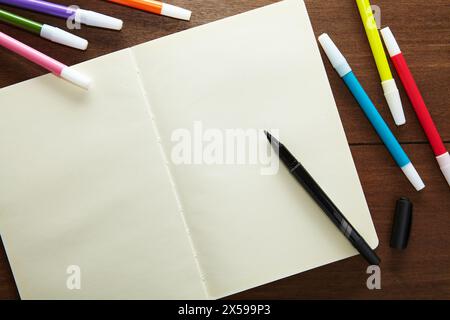 an empty sketchbook opened with a black marker ready to use and a number of coloured markers on the wooden table Stock Photo