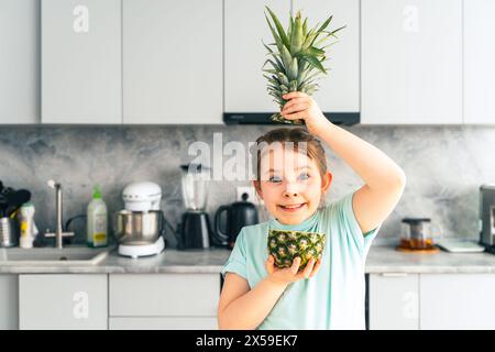 Cute little girl pretending to be a pineapple. Cheerful pre-teen girl playing with pineapple on the background of the kitchen at home. Selected focus. Stock Photo