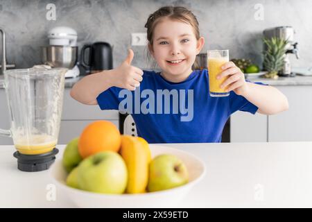 A little girl with freshly squeezed juice in her hand and a thumbs up is sitting at a table with fruit on the background of the kitchen at home. Healt Stock Photo