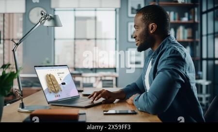 Handsome Black African American Man is Using Laptop Computer with Clothing Online Web Store to Choose and Buy Clothes for New Fresh Look. Beautiful Man Surfing the Internet at Home Living Room. Stock Photo
