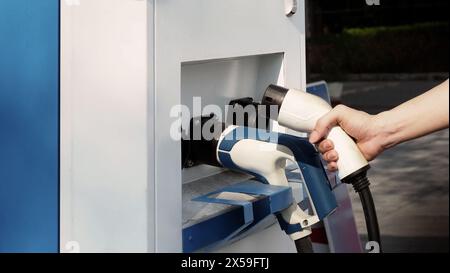 Driver of electric car hold batteries charger plug. Hand holding EV charging plug in front of camera with blurred background of outdoor. Sustainable e Stock Photo