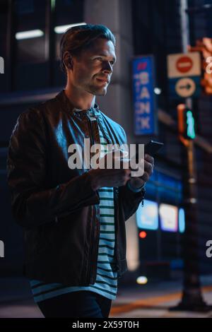 Handsome Young Man Using Smartphone Walking Through Night City Street Full of Neon Lights. Smiling Stylish Blonde Male Using Mobile Phone for Social Media Posting. Vertical Portrait Shot. Stock Photo