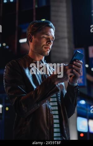 Handsome Young Man Using Smartphone Standing in the Night City Street Full of Neon Lights. Smiling Stylish Blonde Male Using Mobile Phone for Social Media Posting. Vertical Portrait Shot. Stock Photo