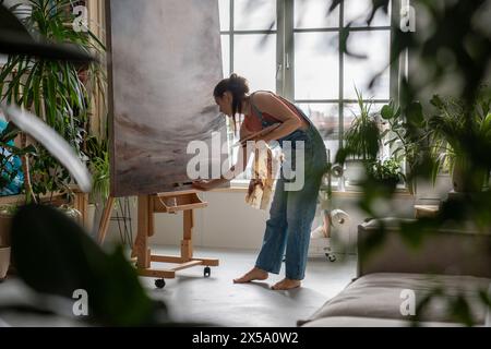 Calm artistic female working on painting in bright creative studio among green plants and flowers. Stock Photo