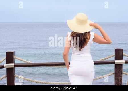A woman in a white dress is standing on a pier overlooking the ocean. She is wearing a straw hat and she is enjoying the view Stock Photo