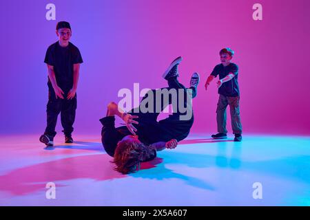 Three young dancers in casual attire perform a vibrant breakdance routine in mixed neon light against vibrant gradient background. Stock Photo