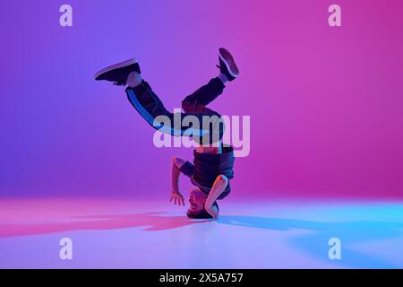 Athletic teenager, break-dancer spinning on head in motion in mixed neon light against vibrant gradient background. Stock Photo