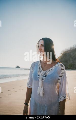 A peaceful young woman in a white dress gazes into the distance on a pristine Costa Rican beach, embracing the tranquility of the shoreline at sunset Stock Photo