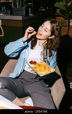 A woman enjoys a tray of golden French fries with a side of ketchup in a relaxed dining setting, embodying a casual and enjoyable mealtime. Stock Photo