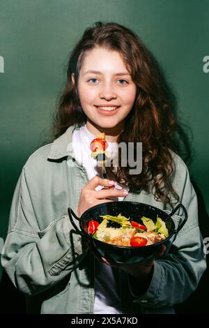 A joyful young woman holding a bowl of Caesar salad, ready to eat a bite of chicken and cherry tomato against a green background Stock Photo