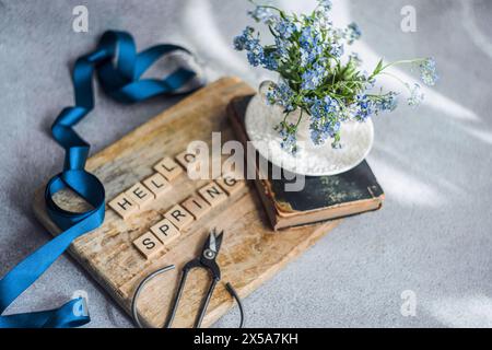 A bouquet of fresh forget-me-not flowers in a white vase, placed on a vintage book with the message HELLO SPRING spelled out in wooden letters, accent Stock Photo