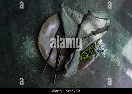 A sophisticated table setting featuring a ceramic plate, dark cutlery, a linen napkin with a decorative ring, and a bouquet of fresh Forget Me Not flo Stock Photo
