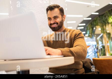 A cheerful man works on his laptop in a modern cafe setting, his smile suggesting productivity and satisfaction with his work Stock Photo
