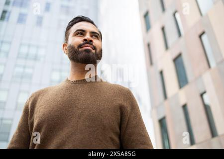 Confident urban man with a beard standing outdoors, looking up thoughtfully Stock Photo