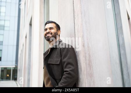 A content man with a beard in a brown coat leaning against an urban building wall, looking upward and smiling Stock Photo