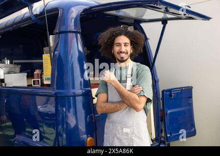 A jovial man with curly hair stands confidently outside his blue food truck, sporting a striped apron and giving a thumbs up Stock Photo