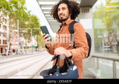 A young man with curly hair, wearing a casual brown jacket, is holding a smartphone while standing with an electric scooter on a city street, possibly Stock Photo