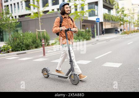 A cheerful young man with a curly hair and beard rides an electric scooter down a city street, wearing a brown jacket and a helmet Stock Photo