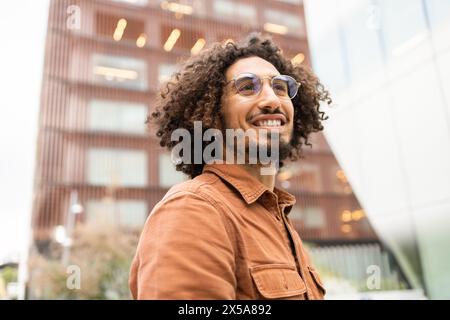 Joyful young man with curly hair rides an electric scooter through an urban area, expressing freedom and happiness against a modern building backdrop Stock Photo