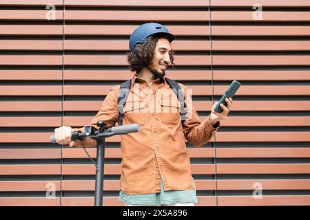 Young man with a helmet, standing with an electric scooter, happily using his smartphone against a striped background Perfectly captures a moment of m Stock Photo