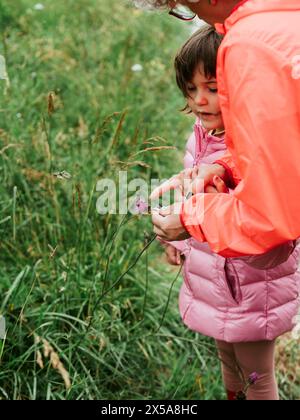 Grandmother in pink jacket spending quality time with her granddaughter outdoors, teaching about plants and flowers. Stock Photo