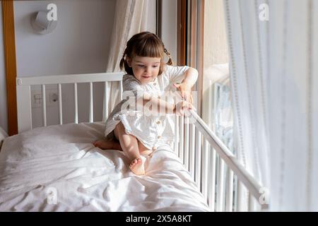 Cheerful toddler girl playing on a bed near a sunny window in a cozy bedroom setting Stock Photo