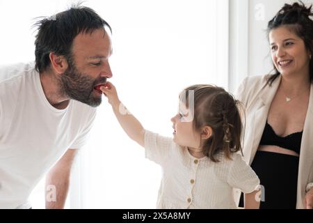 A heartwarming family moment with a toddler feeding her father a snack while her pregnant mother watches with a joyful smile Stock Photo