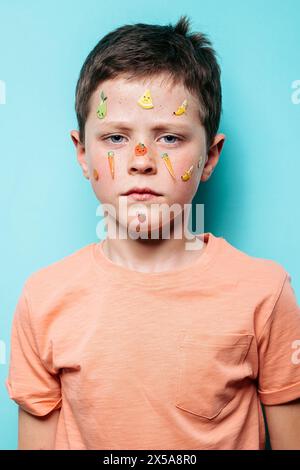 A boy gazes at the camera with a neutral expression, his face adorned with an array of playful fruit-themed stickers against a soft blue backdrop Stock Photo