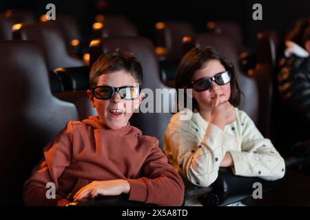 Two kids, a boy and a girl, wearing 3D glasses and watching a movie with intrigued expressions during a summer day at the cinema. Stock Photo