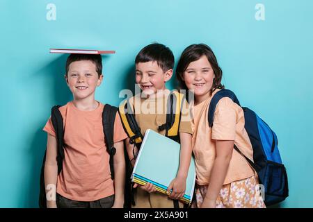 Three cheerful school children, two boys and a girl, stand against a turquoise background. One boy balances a book on his head and smiles at the camer Stock Photo