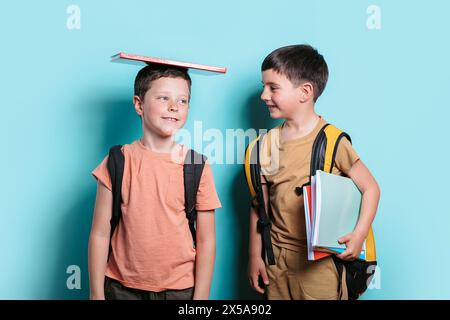 A boy with a book on his head looks away, standing next to his friend who is holding books and looking at him. Stock Photo