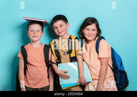 Three cheerful school children, two boys and a girl, stand against a turquoise background. One boy balances a book on his head and smiles at the camer Stock Photo