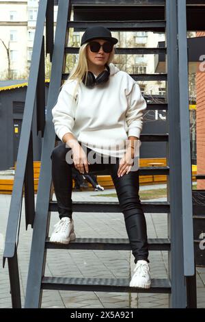 A fashionable blonde lady relaxes on a metal stairway in the city, sporting chic sunglasses and a trendy black hat. Stock Photo