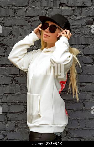 A fashionable blonde woman in sunglasses and a black hat poses confidently against a brick wall backdrop. Stock Photo