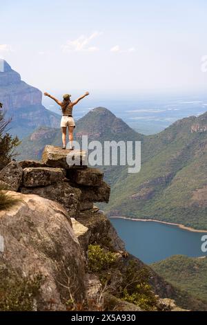 Back view of unrecognizable woman stands triumphantly on a rocky ledge above a verdant valley, with arms outstretched, facing away from the camera, ov Stock Photo