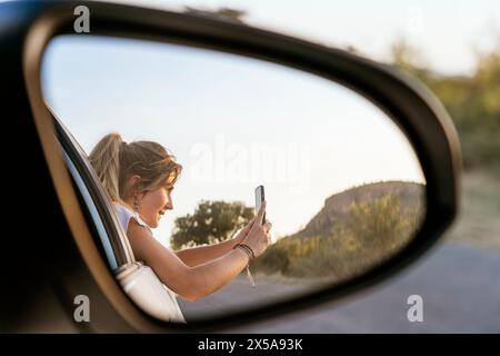 A woman is seen taking a photo of the scenic outdoors with her smartphone, as reflected in a car's side mirror. Stock Photo